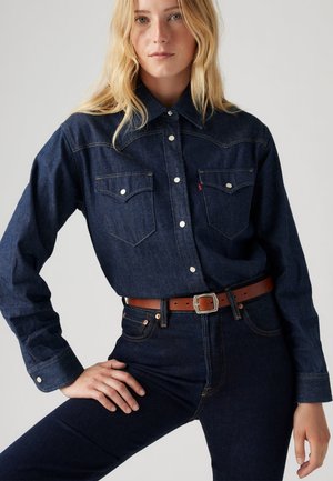 Mørkeblå denimskjorte med to frontlommer, trykknapper og spiss krage, kombinert med mørke jeans og et brunt belte.