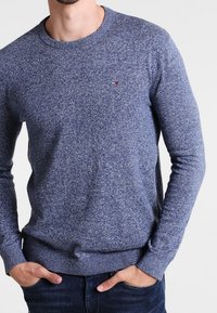 Blauer melierter Strickpullover mit langen Ärmeln, rundem Ausschnitt und geripptem Saum. Kleines Logodetail auf der Brust. Weiche Textur.