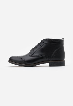 Bottine brogue pour homme en cuir noir avec lacets sur le devant, perforations décoratives et talon empilé sur fond blanc.