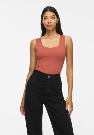 Femme aux longs cheveux foncés portant un haut sans manches couleur rouille et un pantalon noir taille haute sur un fond clair uni.