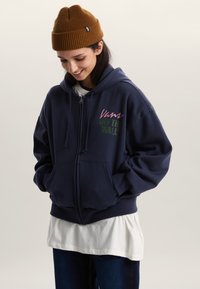 Marineblauwe hoodie met ritssluiting en voorzakken, met roze en groene "Vans Off the Wall"-logo. Gecombineerd met een witte gelaagde shirt eronder.