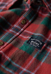 Rood en groene geruite flanellen shirt met bruine knoopsluiting, voorzien van een zwart label met witte tekst. Zachte textuur met zichtbare stiksels.