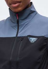 Κοντινό πλάνο ατόμου που φοράει μπλε και μαύρο μπουφάν με ψηλό γιακά και στυλιζαρισμένο λογότυπο με ζωικό πρόσωπο στο στήθος.