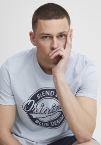 Lyseblå t-skjorte med kort arm og en sirkelformet grafikktrykk som har teksten "Blend in Blue Denim". Glatt bomullstekstur, design med rund hals.