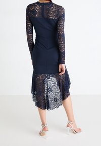 Mörkblå klänning med långa spetsärmar och spettsid, med en figurnära kropp och utsvängt nederdel som framhäver blommiga spettsmönster.