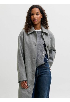 Femme aux cheveux bouclés portant un long manteau gris par-dessus un pull en maille et un jean bleu, debout devant un fond clair uni.