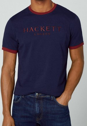 Homme portant un t-shirt bleu marine avec une bordure rouge et le texte "Hackett London", associé à un jean bleu foncé, main dans la poche.