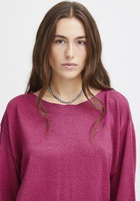 Magenta stickad tröja med bred halsringning och sjunkande axlar, med en mjuk textur. Accenterad med ett silverkedjehalsband.