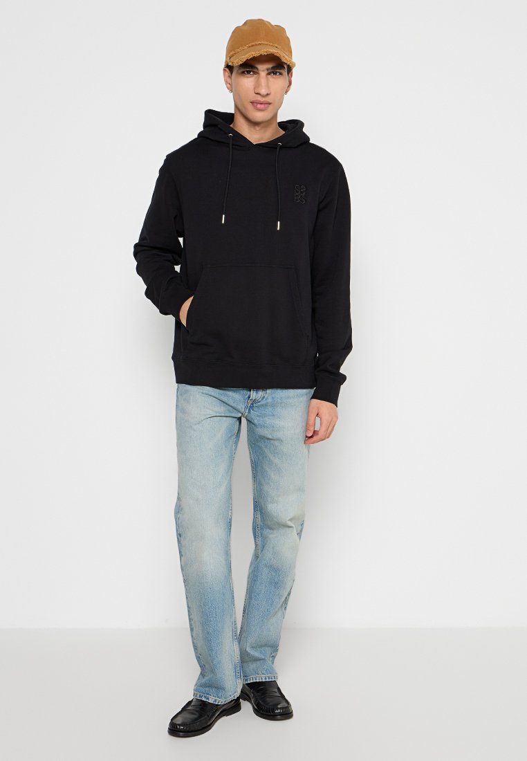 Schwarzer Kapuzenpullover mit einer Fronttasche, Kordelzugkapuze und Logo; kombiniert mit hellblauen Jeans und einer beigen Kappe. Schwarze Schuhe runden das Outfit ab.