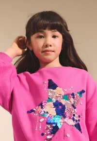 Roza jopa z zvezdnim motivom iz pisanih sequins in perlic, ki prikazuje različne teksture in oblike v živahnih odtenkih.