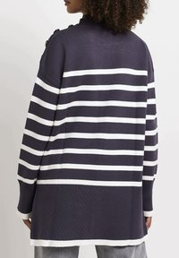 Femme portant un long pull bleu marine à rayures blanches avec des détails de boutons sur l'épaule, vue de dos.