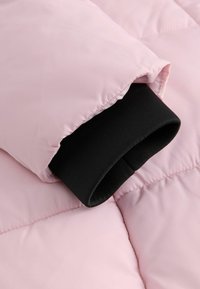 Giacca puffer rosa chiaro con un tessuto esterno liscio, dotata di polsini in maglia nera e dettagli trapuntati per un'ulteriore texture e isolamento.