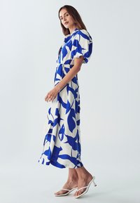 Mėlynai ir baltai abstrakčiai raštuota suknelė, iki kelių, su pūstomis rankovėmis, tekstūrinė medžiaga ir deranti juosta juosmens srityje. Su baltais aukštakulniais.