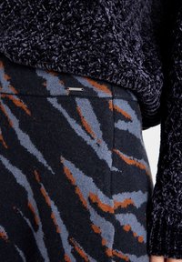 Pull en tricot bleu foncé avec une texture duveteuse. En dessous, un pantalon à motifs présente des rayures orange vif et bleu clair sur un fond sombre, avec un accent métallique.