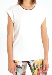 Vit bomullst-shirt med uppvikt ärm och marinblå halsringning, parat med färgglada leggings med fågelmönster och livliga färger.