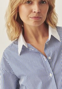 Camicia a righe blu e bianche con colletto bianco. Presenta righe verticali e un tessuto liscio, che mostra una vestibilità rilassata.