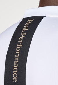 Hvit treningsskjorte med svart sidestripe. Stripen har "PeakPerformance" i gullbokstaver, som skaper en kontrasterende design detalj.