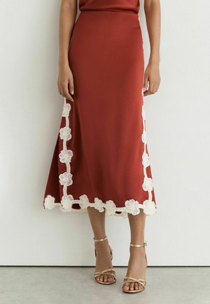 Jupe midi couleur rouille avec un bord en dentelle florale blanche sur les côtés et l’ourlet, portée avec des sandales dorées à bride cheville sur un sol clair.