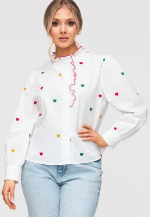 Kvinde iført en hvid bluse med farverige hjerteprint og lyserød flæsekant, kombineret med lyseblå jeans, poserer med den ene hånd på hoften.