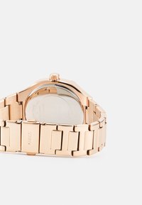 Montre-bracelet ton or rose avec un bracelet en métal, boîtier rond et finition polie. Elle présente un fond en verre visible et un logo gravé.