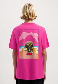 Roze katoenen T-shirt met een grote cartoonafbeelding op de achterkant, waarop een figuur met een schild en een kleurrijke strandachtergrond is afgebeeld.