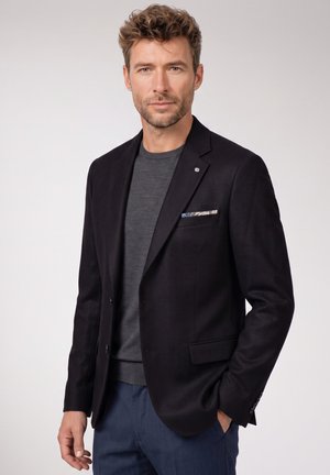 Man with curly hair wearing a black blazer, gray sweater, and blue pants, standing with one hand in pocket against plain background.