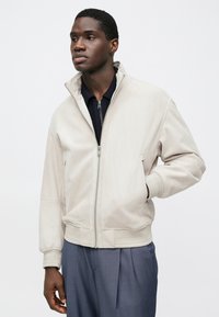 Veste bomber beige clair avec un col montant, une fermeture éclair à l'avant et des poignets côtelés. Texture lisse, associée à une chemise bleu marine et un pantalon coupé sur mesure.
