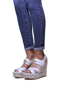 Bílé klínové sandály s více pásky, zlatým kruhovým akcentem a strukturovanou podrážkou, v kombinaci s ohrnutými tmavými džíny.