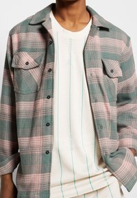 Rutigt skjorta med knappar i teal- och rosa nyanser med två bröstfickor. Lager på en texturerad vit mesh-topp med turkosa ränder.