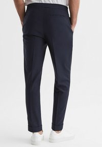 Pantalones marinos en un ajuste entallado con un frontal plano y puños enrollados. Presenta un bolsillo trasero y cinturilla de tela. Combinados con zapatillas blancas.