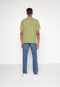 Olijfgroene katoenen T-shirt met korte mouwen, ontspannen pasvorm, gecombineerd met blauwe denim jeans en witte sneakers, gezien van achteren.
