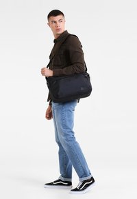 Borsa a tracolla nera in nylon con zip superiore, tracolla regolabile e logo frontale. Modello che indossa pantaloni di jeans casual e sneakers.