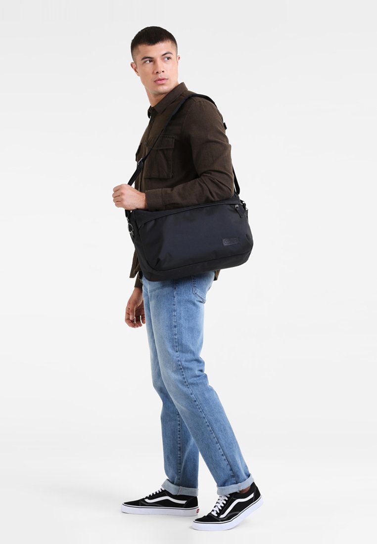 Borsa a tracolla nera in nylon con zip superiore, tracolla regolabile e logo frontale. Modello che indossa pantaloni di jeans casual e sneakers.