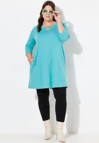 Aqua tuniekjurk met ¾ mouwen, een V-hals met kruislingse banden en zakken, gecombineerd met zwarte leggings en crèmekleurige enkelboots.
