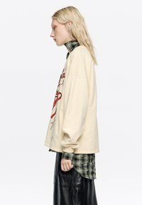 Veľký krémový sveter s červenou grafikou a detailmi hviezd cez zelenú károvanú košeľu, doplnený lesklou čiernou sukňou. Dlhé rukávy.