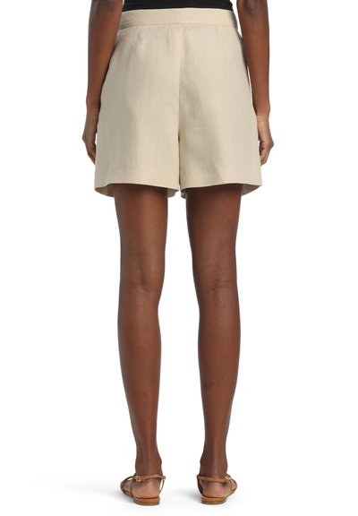 Set bagfra af en person iført beige højtaljede shorts kombineret med brune flade sandaler, stående foran en hvid baggrund.