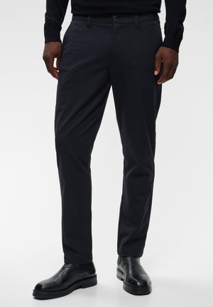 Mann trägt schwarze, maßgeschneiderte Hose, schwarzes langärmeliges Hemd und schwarze Lederschuhe und steht auf einem schlichten hellen Hintergrund.