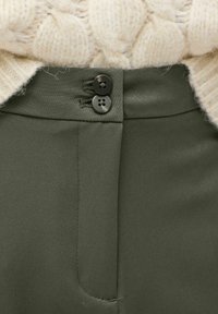 Olijfgroene broeken van een gladde stof, met twee zwarte knopen in de taille en nette stiksels, gedeeltelijk bedekt door een crèmekleurige kabeltrui.