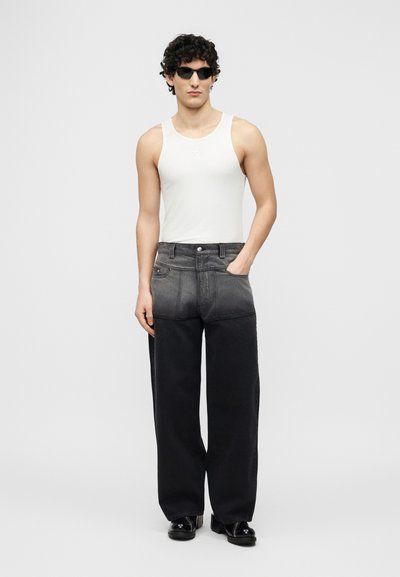 Άνδρας με σγουρά μαλλιά που φοράει μαύρα γυαλιά ηλίου, λευκό αμάνικο τοπ, φαρδιά παντελόνια με γκρι προς μαύρο βαθμιαίο χρώμα και μαύρα παπούτσια, στέκεται με ένα χέρι στην τσέπη.