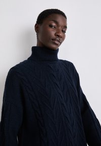 Ναυτικό πουλόβερ με πλέξη καλτσοποίησης και ψηλό rib κολάρο, με υφές από φύλλα και φαρδιά μανίκια, κατασκευασμένο από μαλακό, παχύ υλικό.