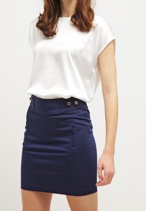 Mujer que lleva una blusa blanca de manga corta metida en una falda azul marino ajustada con detalles de botones en la cintura.