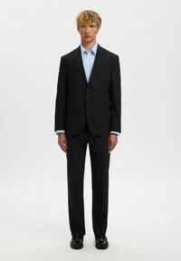 Zwarte pak met een getailleerde blazer en rechte broek, voorzien van een notch lapel, enkele knoopsluiting en een lichtblauw overhemd met kraag.
