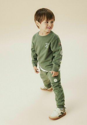 Felpa verde in cotone con grafica bianca e toppe sulle maniche, abbinata a pantaloni coordinati. Presenta una maglietta bianca sotto e sneaker beige.