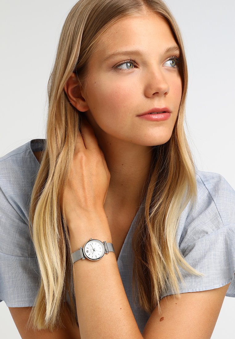 Srebrna mrežasta wristwatch z belo številčnico, črnimi številkami in tankimi kazalci. Nosi se na zapestju, kjer je delno vidna dolga blond las.