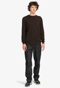 Dunkelbrauner Strickpullover mit Rundhalsausschnitt, langen Ärmeln und gerippten Bündchen, kombiniert mit dunkelgrauen Jeans und schwarzen Sportschuhen.