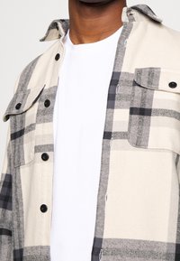 Beżowo-szara koszula w kratę z zapięciem na guziki, z kieszeniami na piersi i białą koszulą pod spodem. Miękki materiał o luźnym kroju.