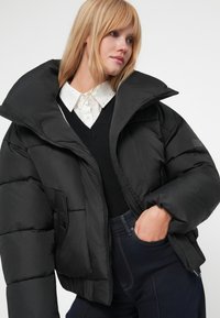 Jachetă puffer neagră cu guler înalt, având un model matlasat și închidere cu fermoar. Purtați peste un pulover negru și o cămașă albă cu guler.