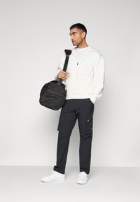 Veste à capuche blanche zippée, pantalon cargo noir et baskets blanches. Portant un sac noir texturé avec une bandoulière, de face.