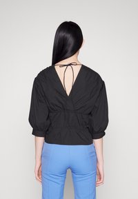 Blouse noire texturée avec un profond décolleté en V dans le dos et une fermeture à nouer, présentant des manches bouffantes et une taille ajustée. Associée à un pantalon bleu clair.