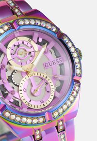 Orologio Guess con cassa metallic rainbow, quadrante rosa con ingranaggi, lancette dorate e lunetta decorata con cristalli. Forma rotonda, design elegante.